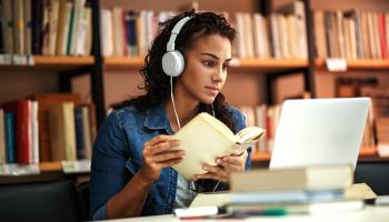 A grad student consults a book while taking an online course on her laptop.